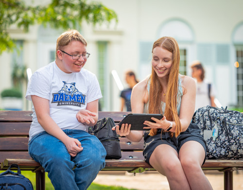 Two students sitting on a bench smiling. One student is holding a table and showing something to the other student.