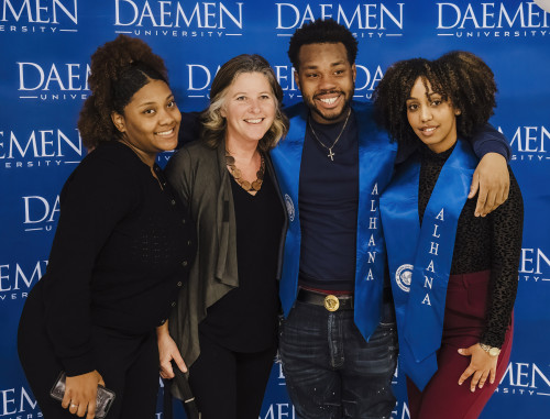 Four people standing in front of Daemen sign; two student wearing ALHANA sashes 
