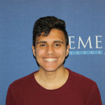 Mateo, a medium skin-toned young man stands in front of a Daemen College wall. He has dark brown hair, dark eyes, a maroon shirt, and is smiling wide.