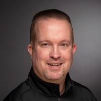 Man with a brush cut wearing a black shirt in front of a gray backdrop