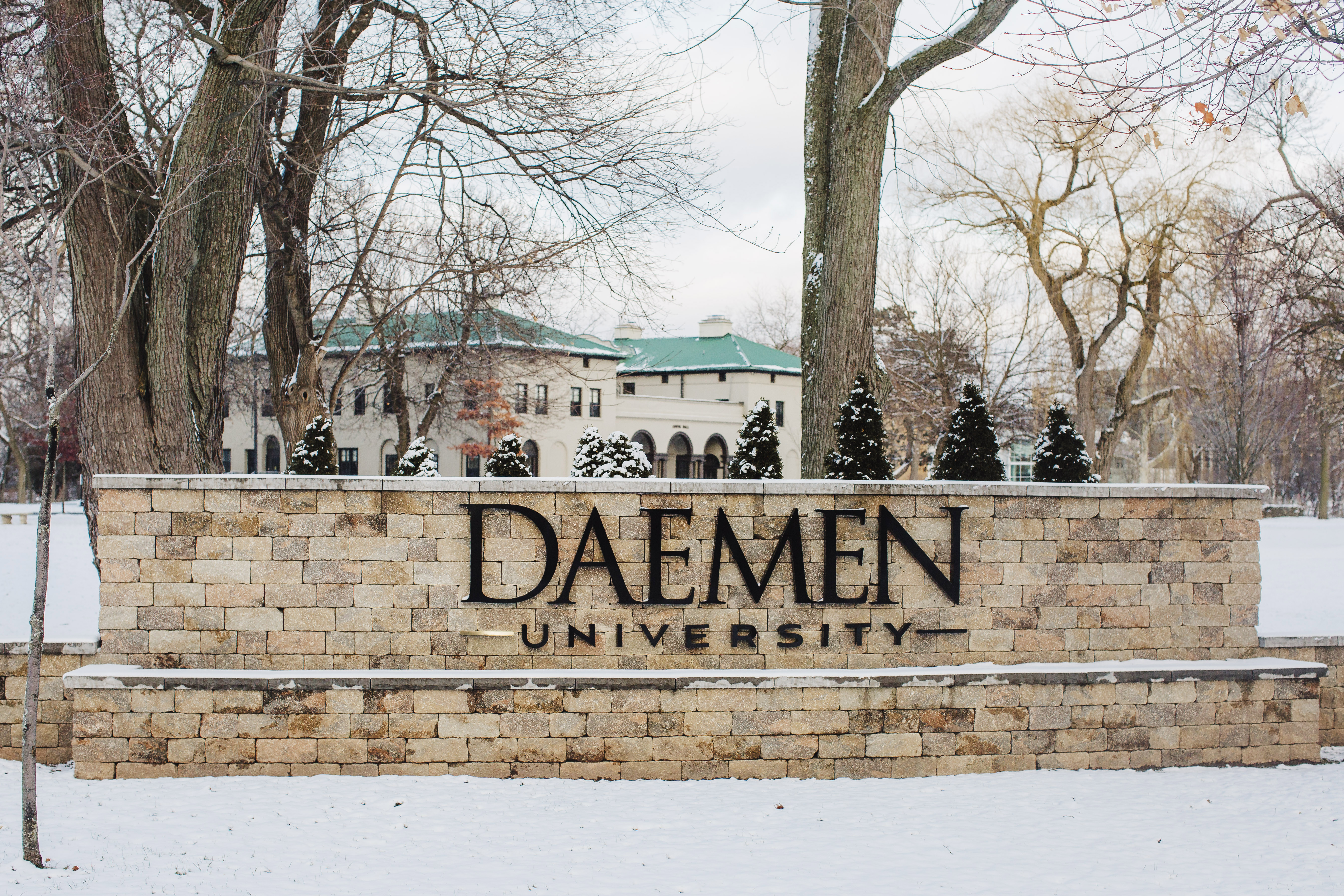 Stone wall with Daemen University letters on it and snow, bare trees and a campus building in the background