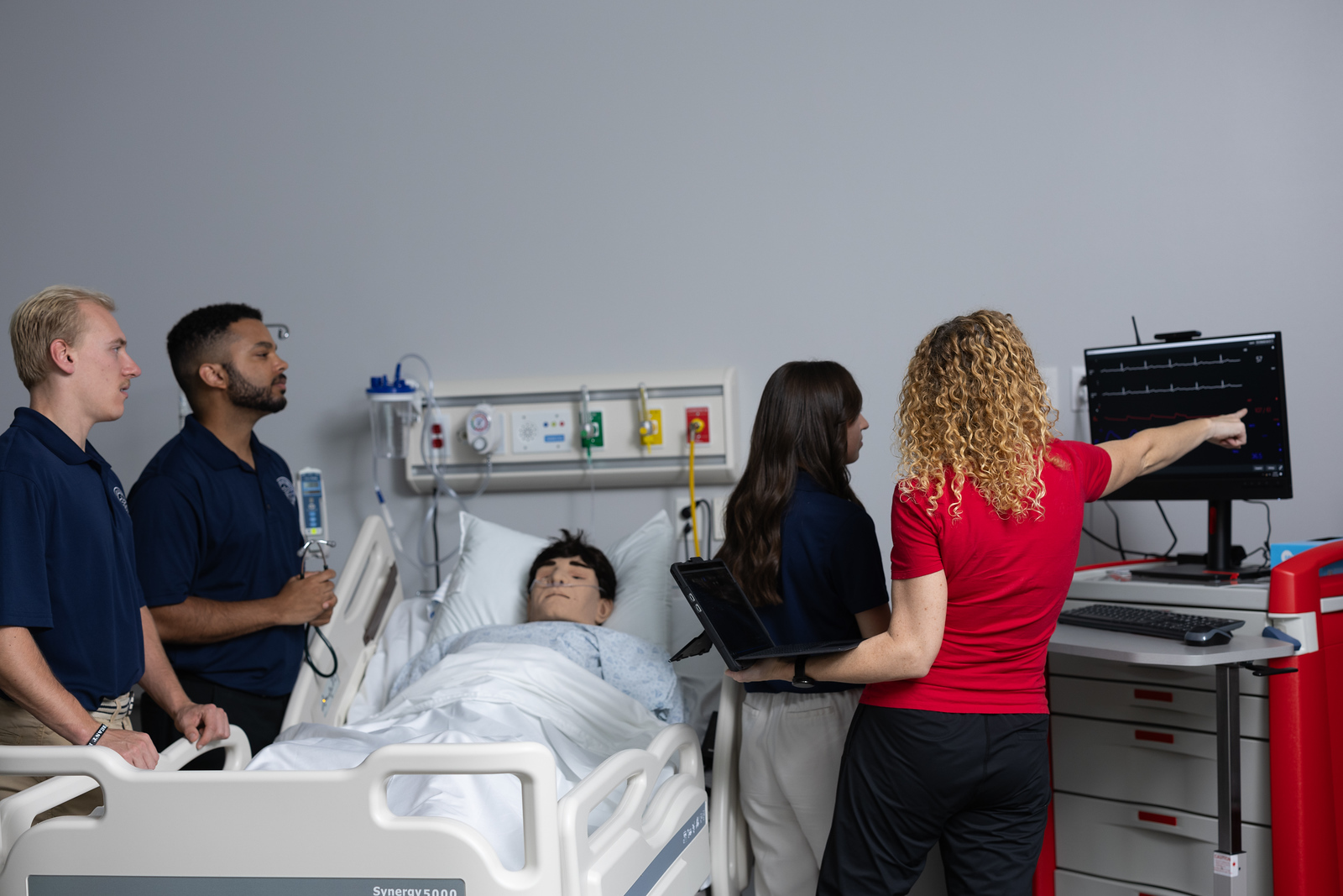 Students standing around a bed with a medical mannequin in it looking at a monitor