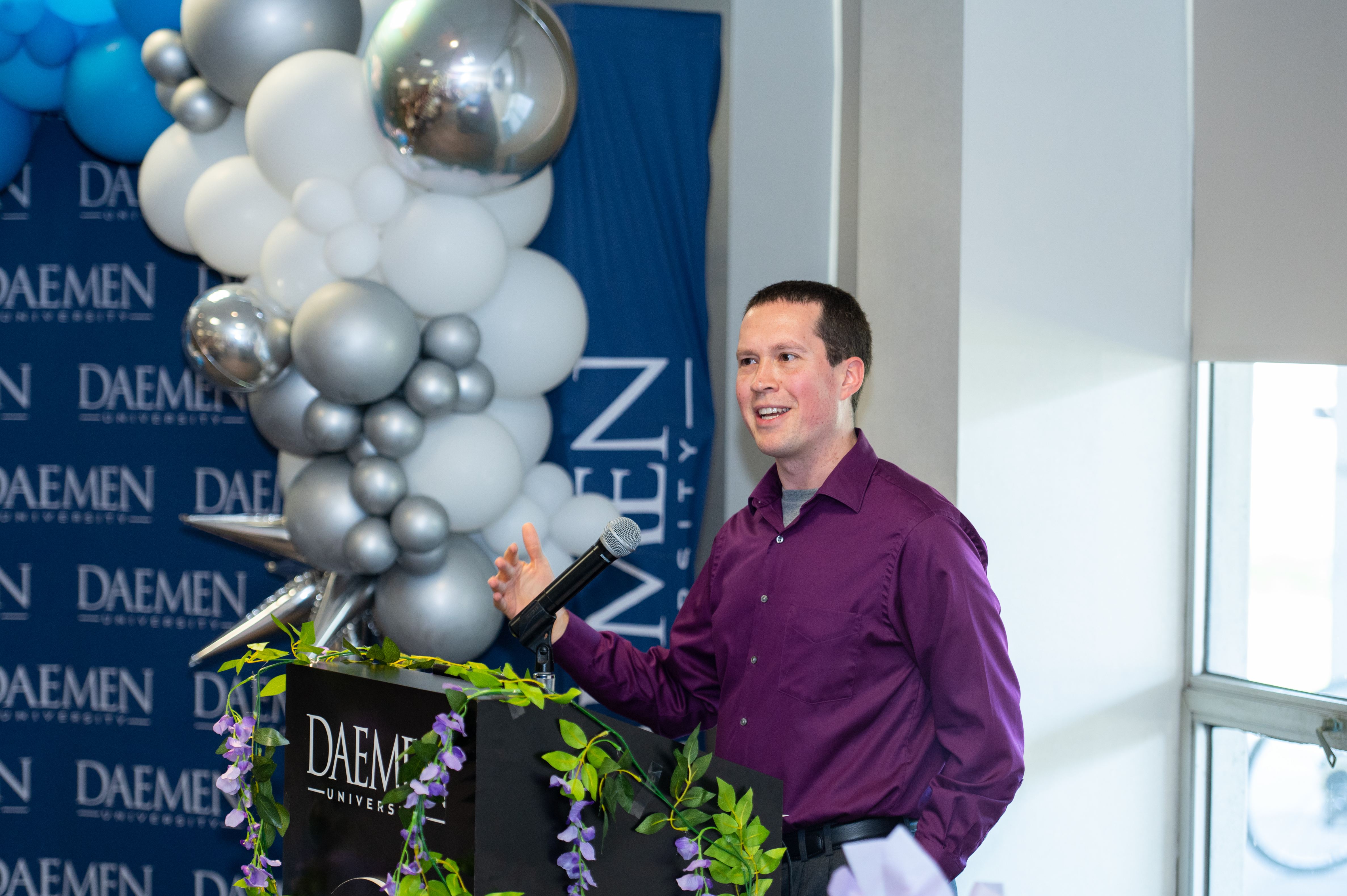 Keynote speaker, Dr. Kurt Blankschaen, wearing a purple button up shirt and standing in front of a podium, delivers a moving address to the graduates and audience