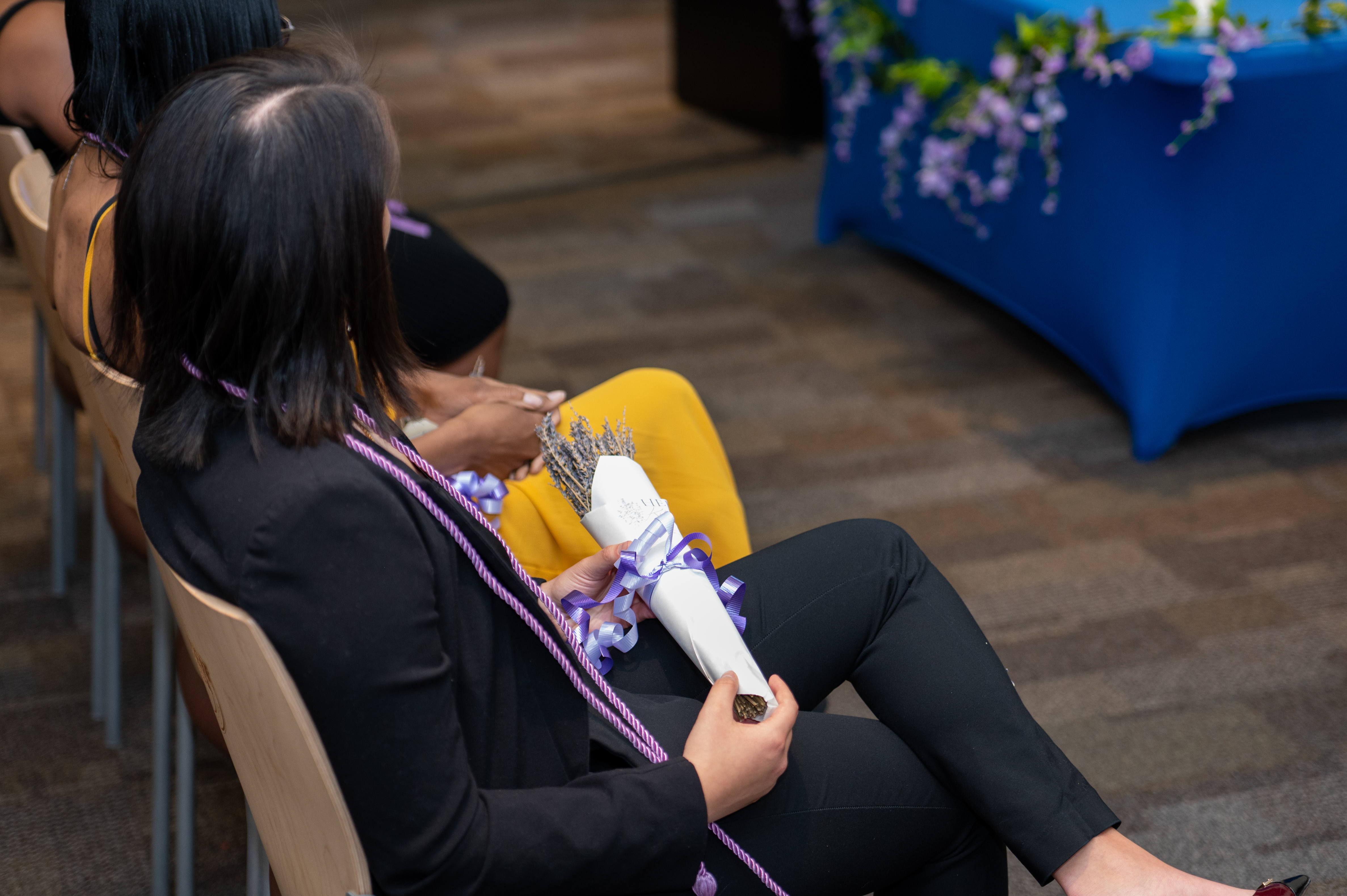 Graduate wearing black suit seated viewing the ceremony holding bouquet of lavendar