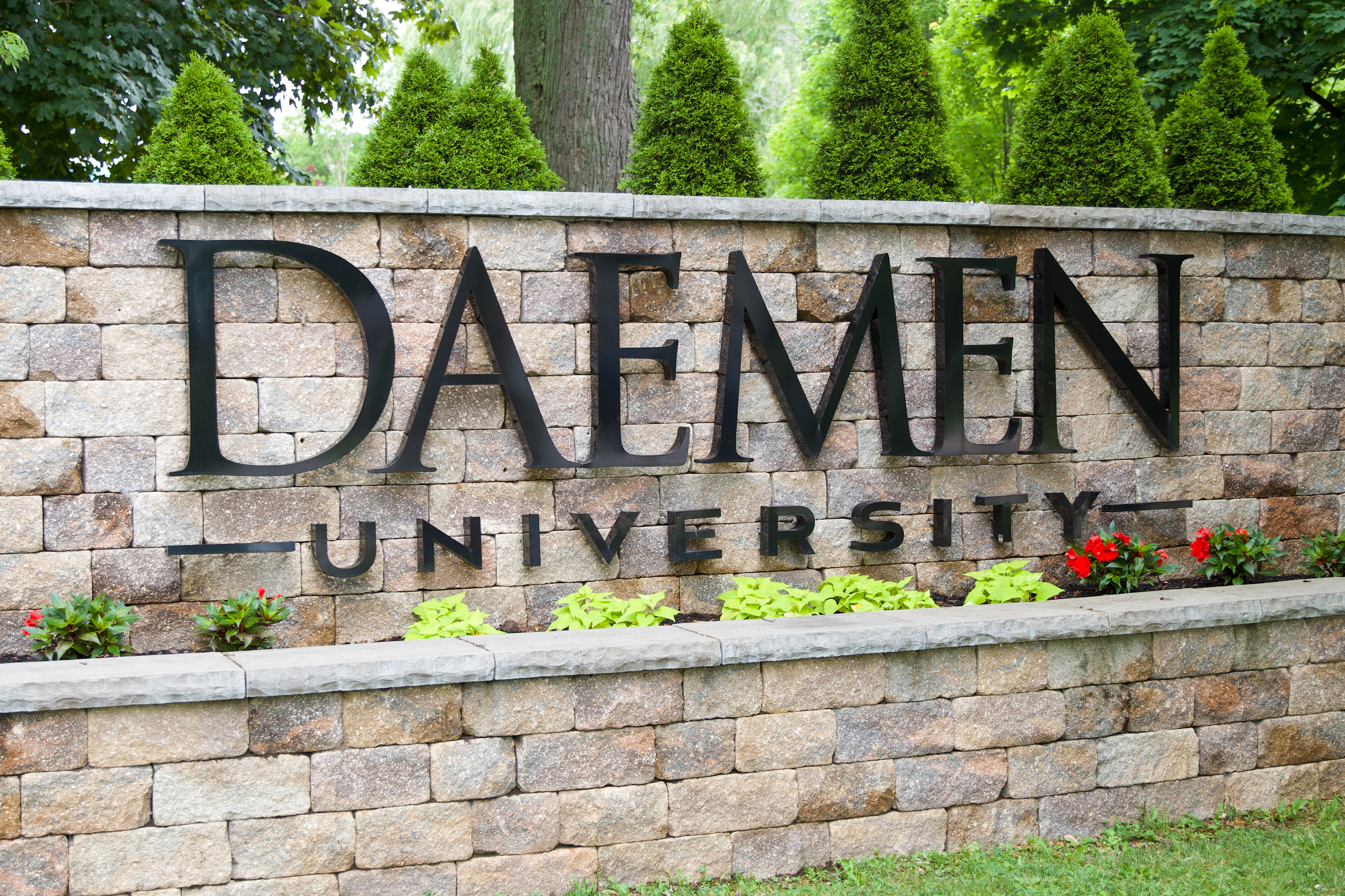 Stone wall with Daemen University sign on it. Flowers are planted underneath the sign and trees are behind the wall. 