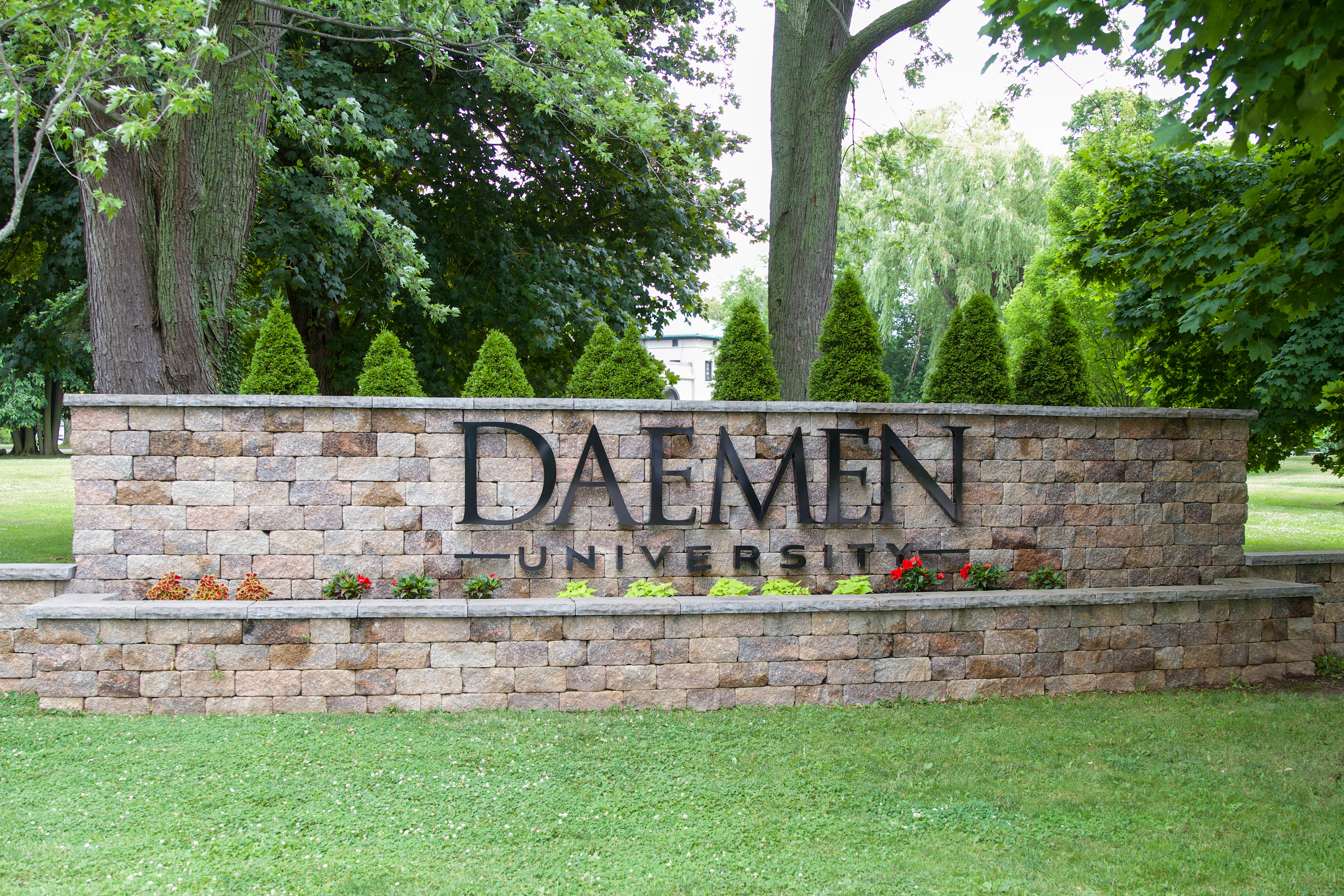 Stone wall with Daemen University metal letters on it, surrounded by grass, trees and flowers