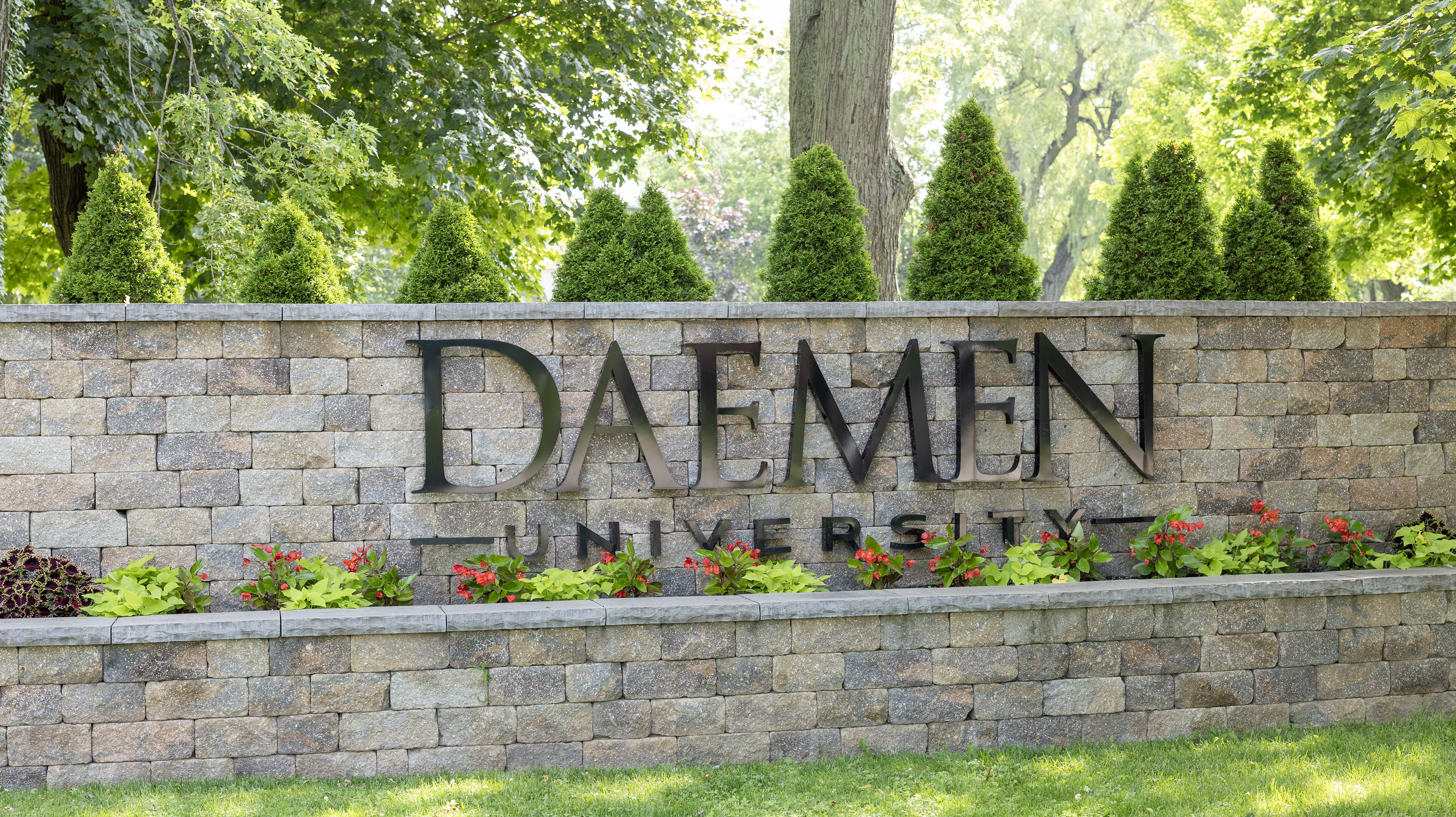 Stone wall with Daemen University sign on it. Flowers are planted underneath the sign and trees are behind the wall. 