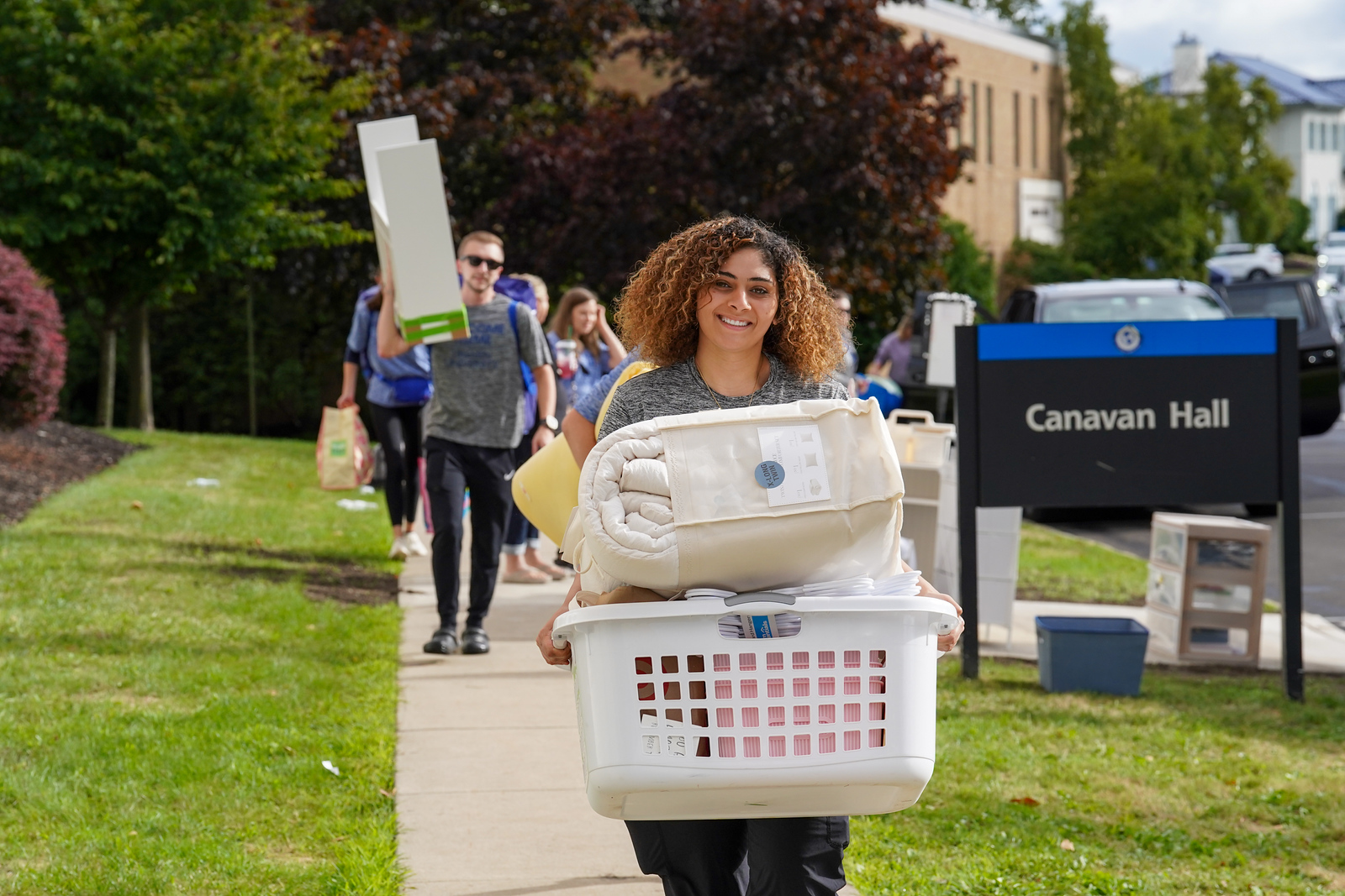 Woman carrying a laundry basket filled with items walking toward residence hall on Daemen campus. Several other people behind her carrying items with grass, cars and buildings in the background