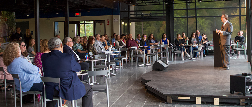 President Olson standing on stage in Daemen Hall atrium speaking to a crown