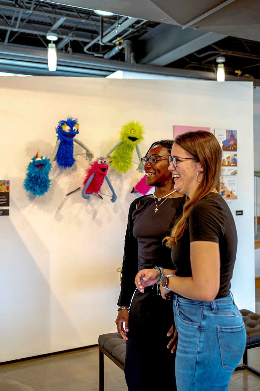 Student in Daemen art gallery, puppets hanging on wall behind them