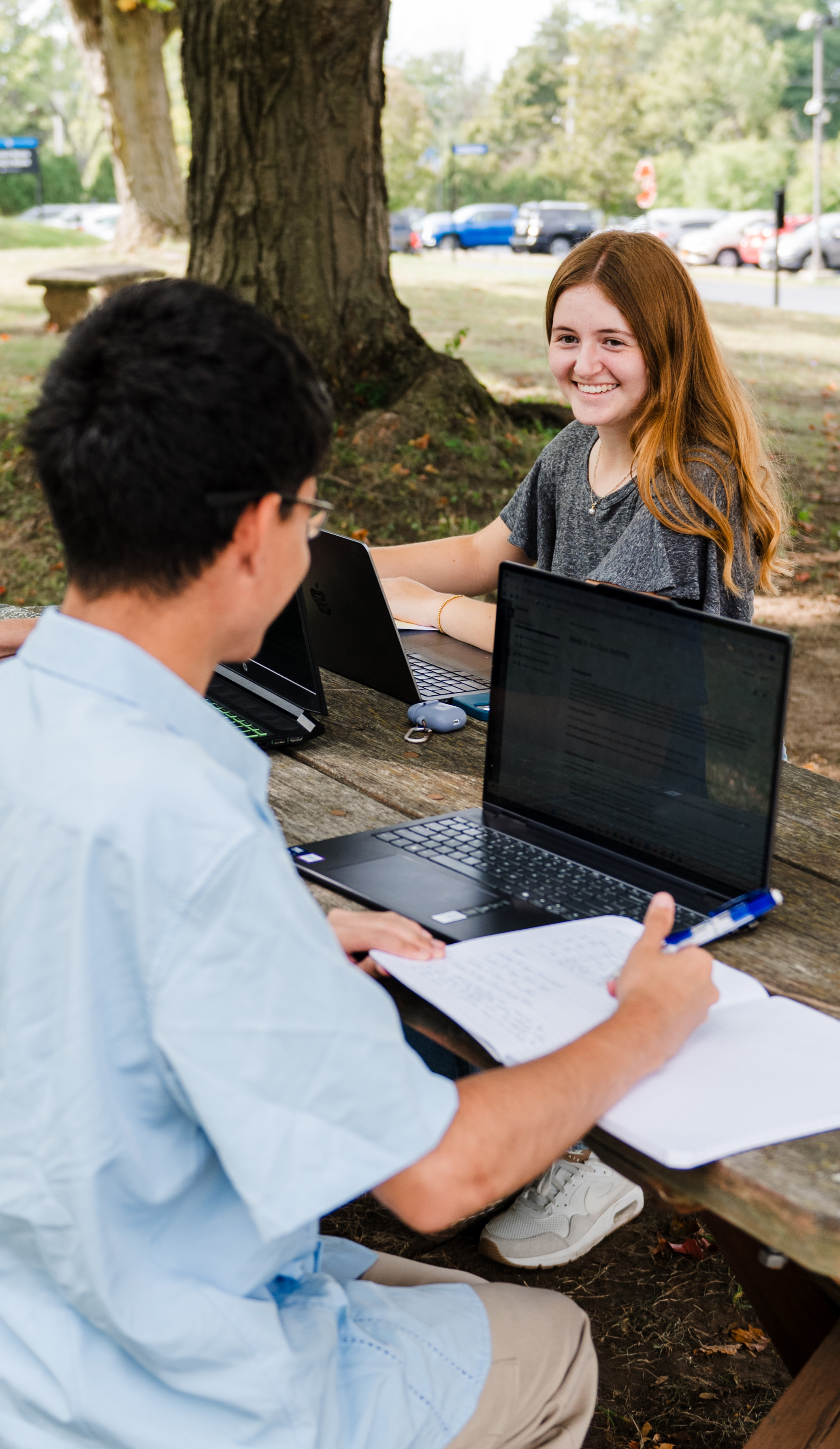 Students sitting outside at a picnic table with laptops and notepads