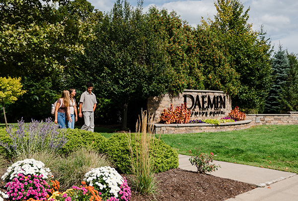 Students walking by stone Daemen sign and wall at front of campus