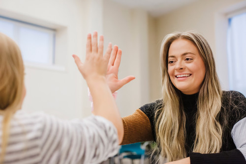 Woman smiling and giving a high-five to another person across a table in a bright indoor setting, suggesting a supportive or therapeutic interaction.