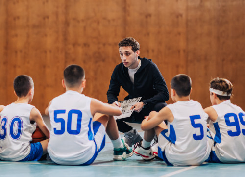 Coach kneeling down showing sitting underage baskball team a play on a dry erase board