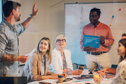 People around an office table one person with their hand up while another holds a slide and is presenting