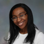 Kimberly is a young black woman smiling for the camera in front of a gray background. She is wearing glasses, a white shirt, and her hair is black and shoulder length.
