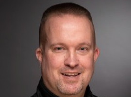 Man with a brush cut wearing a black shirt in front of a gray backdrop