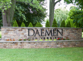 Stone wall with Daemen University metal letters on it, surrounded by grass, trees and flowers