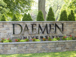 Stone wall with Daemen University sign on it. Flowers are planted underneath the sign and trees are behind the wall. 