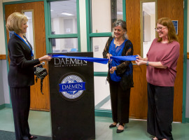 Two women holding a blue ribbon while another woman cuts it with oversized scissors