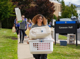 Woman carrying a laundry basket filled with items walking toward residence hall on Daemen campus. Several other people behind her carrying items with grass, cars and buildings in the background