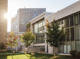 Contemporary building with long windows and gray brick with green grass, trees and a sidewalk in front of it