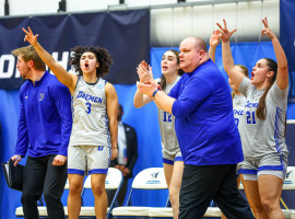 Women's college basketball players standing on the side of the court while the coach claps and walks past them